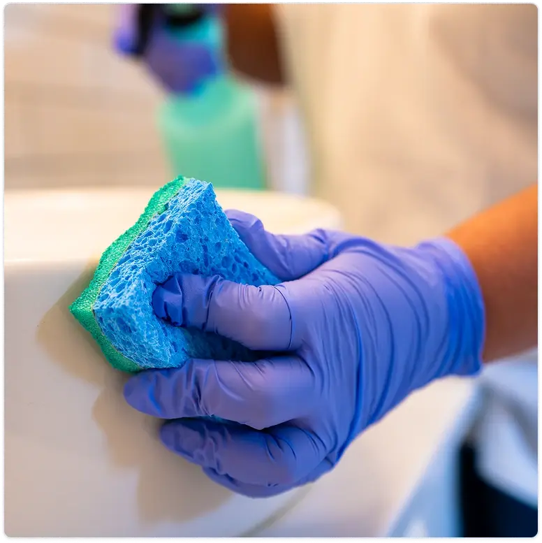Cleaner wearing blue gloves wiping a white bathroom sink with a green cloth